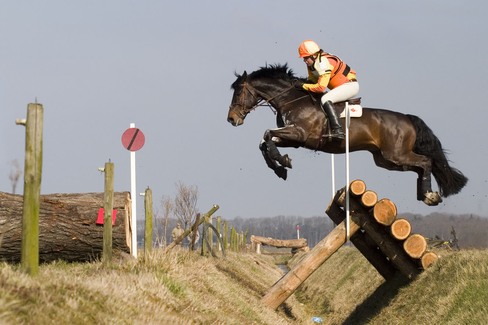 A rider in an orange vest on a dark bay horse jumping a log obstacle built over a ditch on a cross-country course